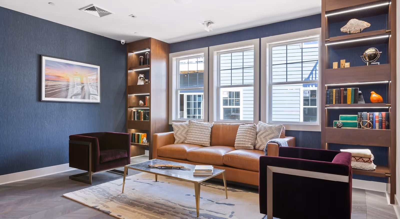 A modern living room with a brown leather sofa adorned with three patterned pillows, two dark purple armchairs, a rectangular coffee table with books and decorative items, built-in wooden shelves with books and decor, and three large windows letting in natural light.