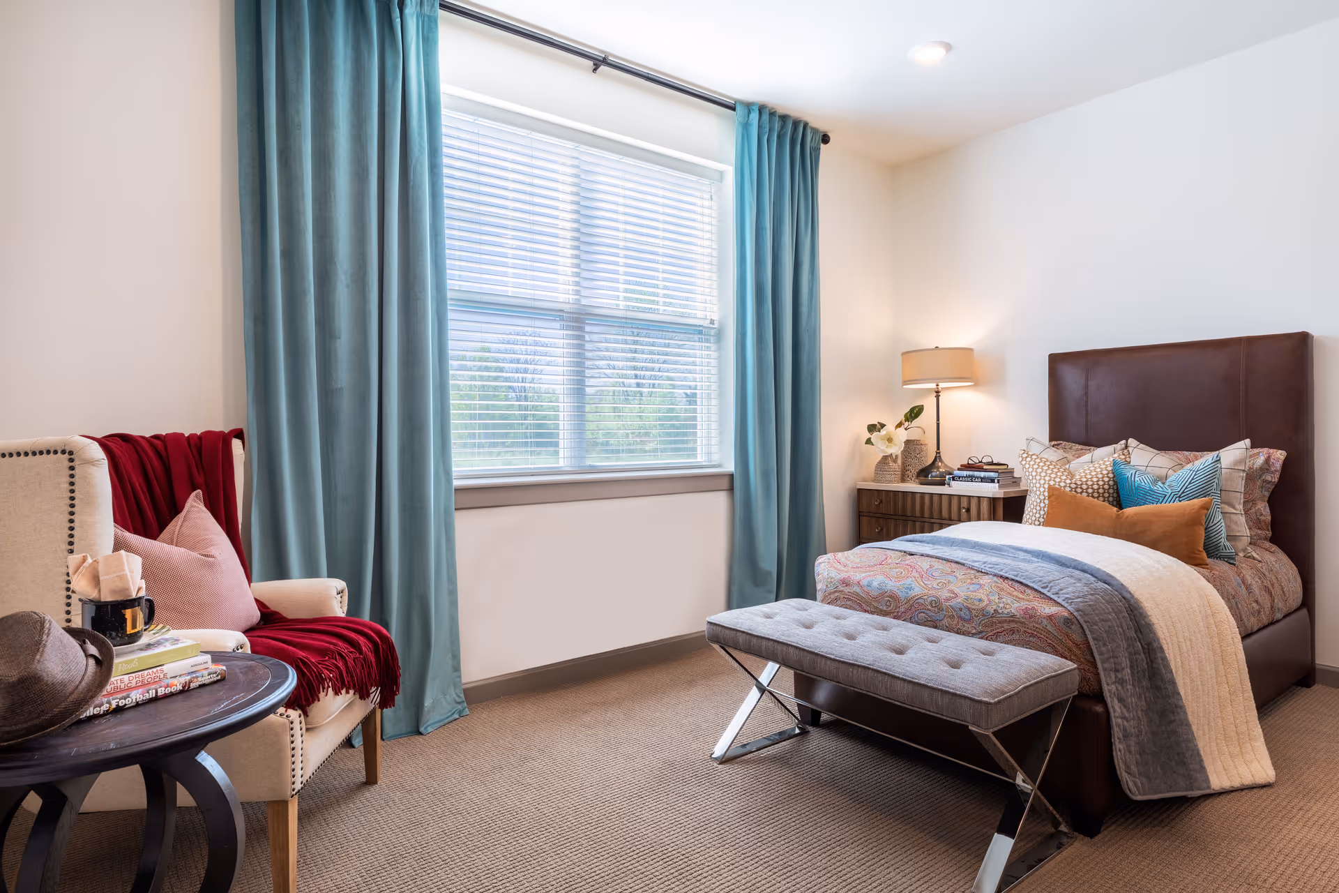 A cozy bedroom with a single bed featuring a brown leather headboard, colorful patterned bedding, and multiple pillows. A gray cushioned bench is placed at the foot of the bed. To the right of the bed is a wooden nightstand with a lamp, books, and a small plant. On the left side of the room, there is a beige armchair with a red throw blanket and a pink pillow, next to a round wooden side table holding a hat, a mug, and some books. The room has a large window with blue curtains and white blinds, and beige carpeting.
