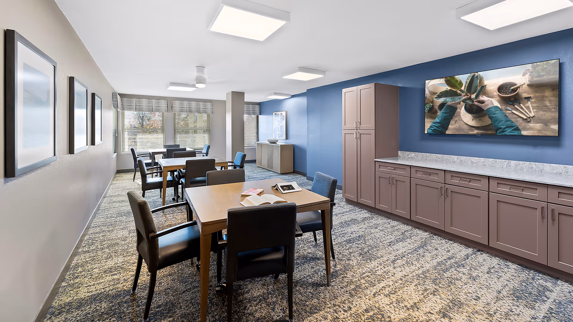 A well-lit common area with multiple tables and chairs arranged for seating. The room has a blue accent wall with built-in cabinets and a countertop. A large flat-screen TV mounted on the wall displays a close-up image of hands tending to a potted plant. The opposite wall features framed artwork, and large windows with blinds allow natural light to fill the space.