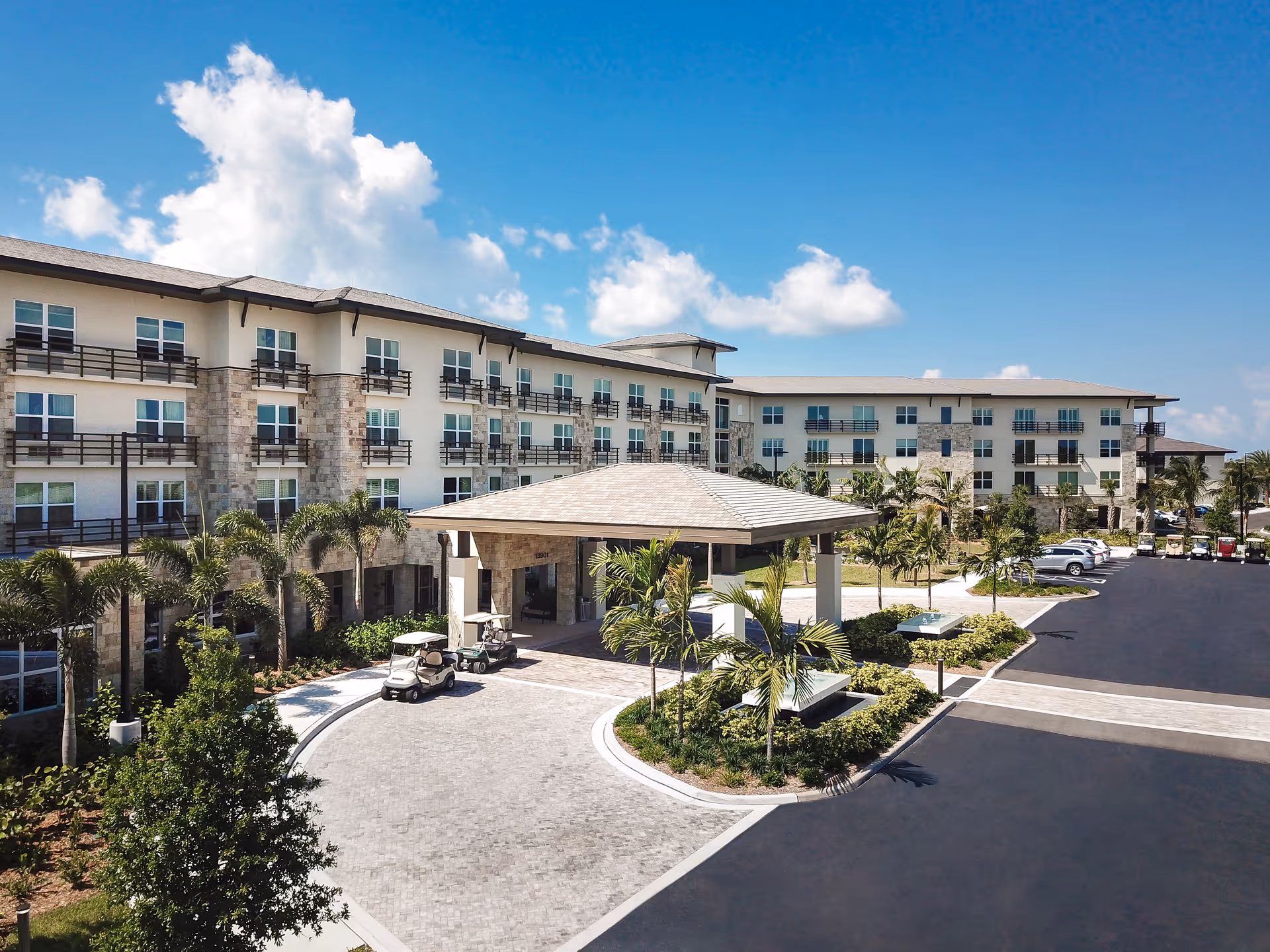 Exterior view of a large senior living facility building with multiple floors, balconies, palm trees, a covered entrance with golf carts parked nearby, and a spacious parking lot under a blue sky with scattered clouds.