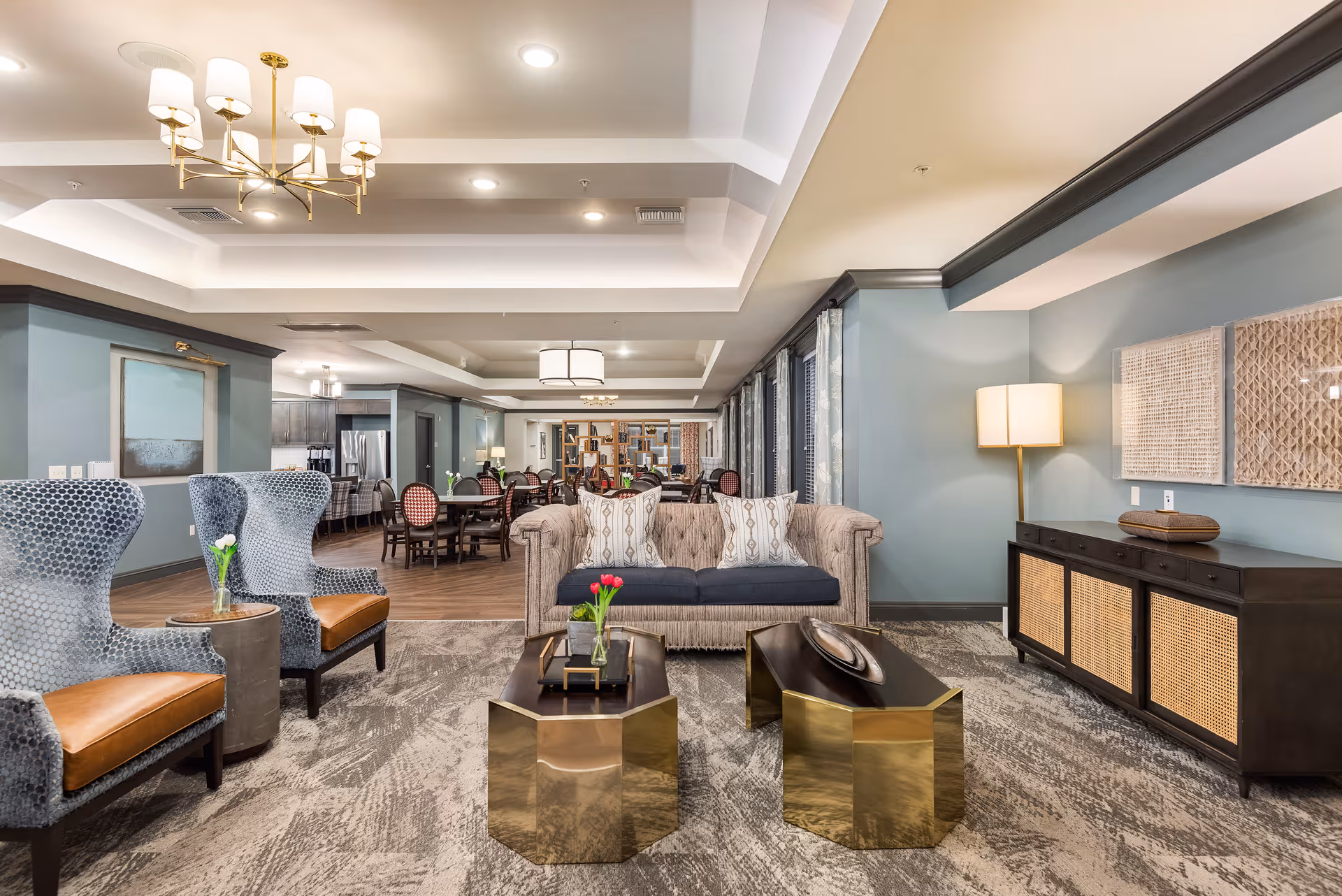 A spacious and well-lit senior living common area with a beige sofa, two blue patterned armchairs with brown cushions, two gold hexagonal coffee tables with decorative items, a sideboard with a lamp and wall art, and a dining area with tables and chairs in the background. The walls are painted light blue and the ceiling has recessed lighting and a chandelier.