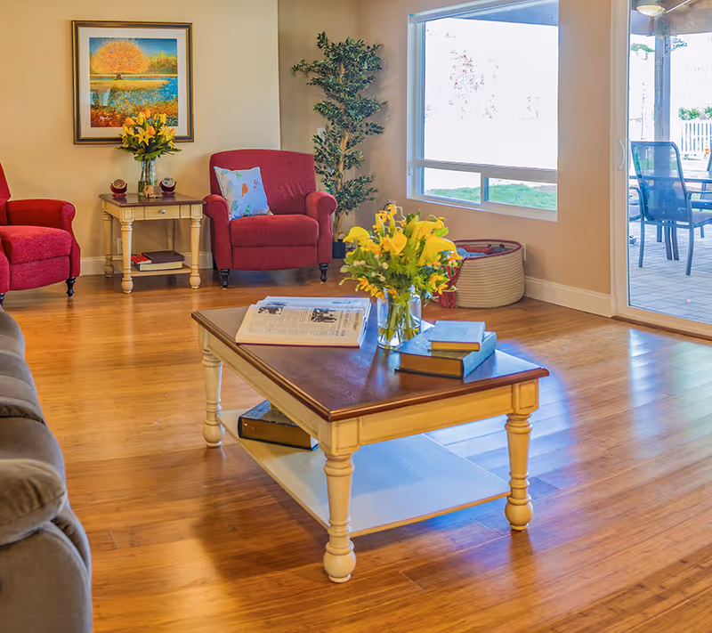 A cozy living room with wooden flooring featuring a wooden coffee table with books and a vase of yellow flowers. There are two red armchairs with a small side table between them holding a vase with flowers and decorative items. A large window and glass door provide a view of an outdoor patio with chairs. A framed painting and a potted plant decorate the room.