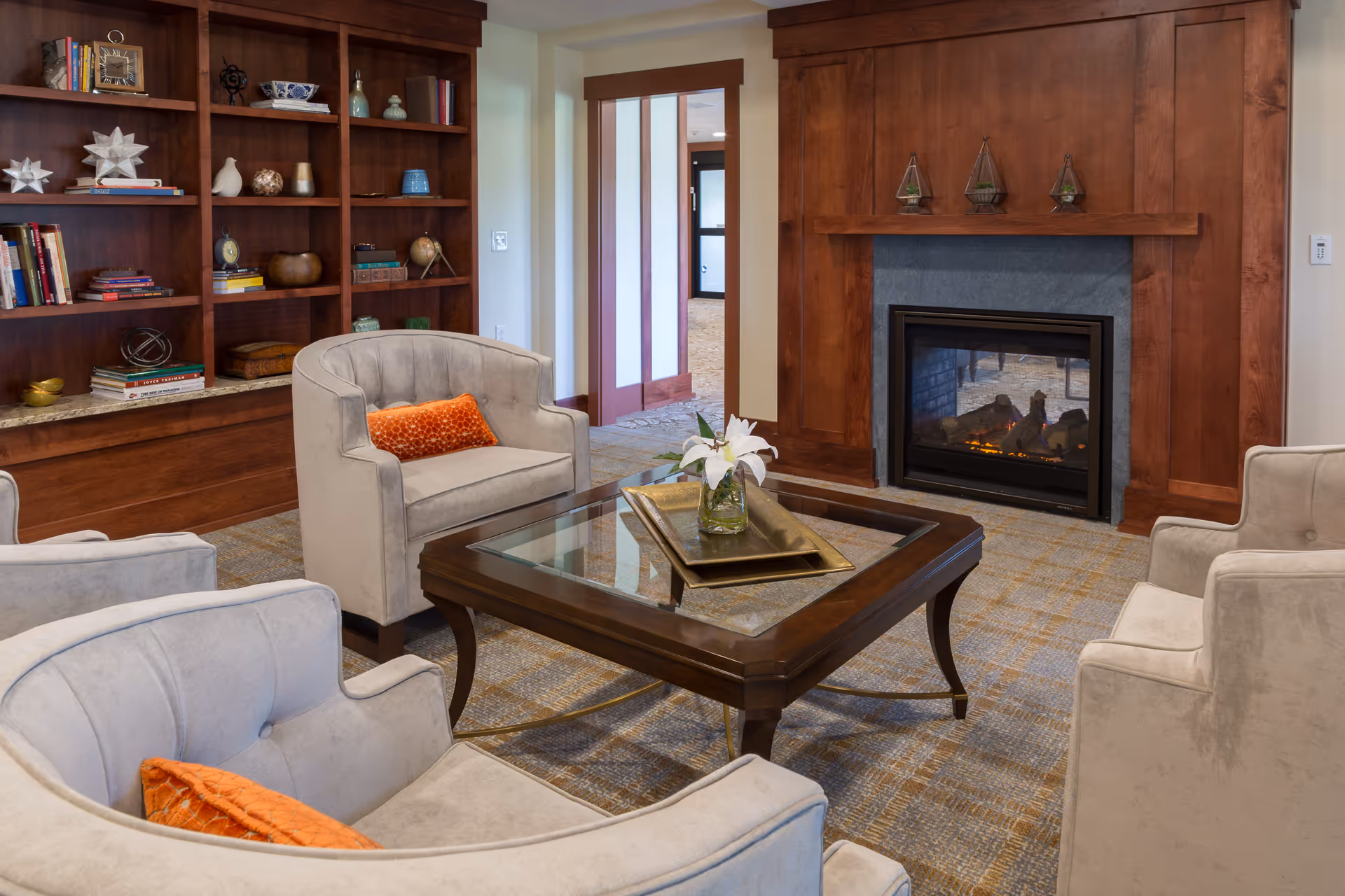 A cozy living room area with four beige armchairs, each with an orange accent pillow, arranged around a wooden coffee table with a glass top. On the table is a decorative tray with a vase holding white flowers. Behind the seating area is a wooden built-in bookshelf filled with books and decorative items. To the right is a wooden fireplace with a lit fire and three small glass terrariums on the mantel.