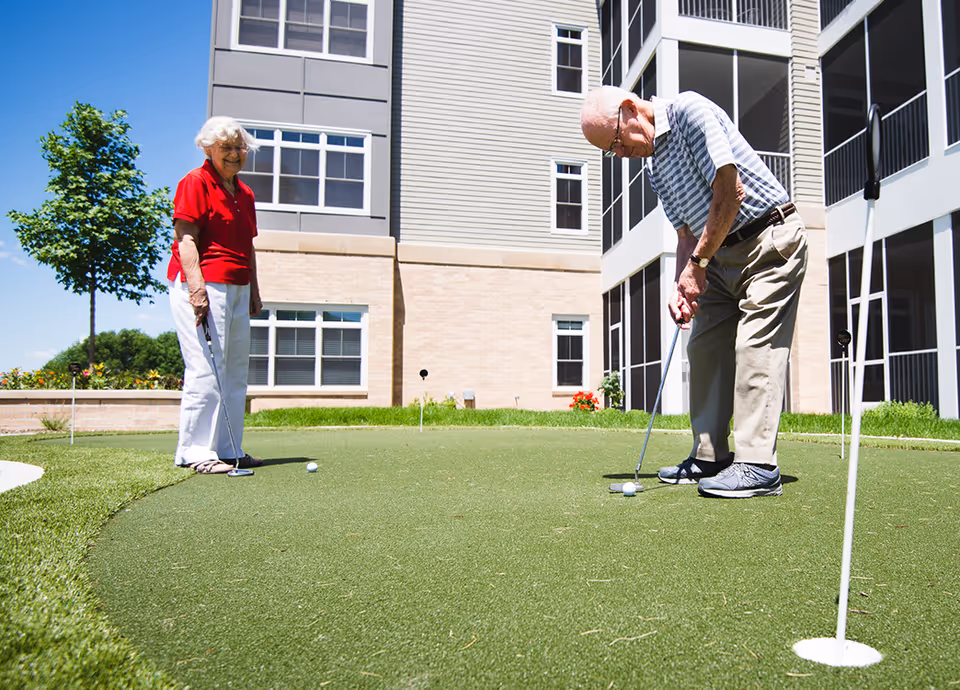 Two seniors playing on a small outdoor putting green next to a multi-story senior living building.