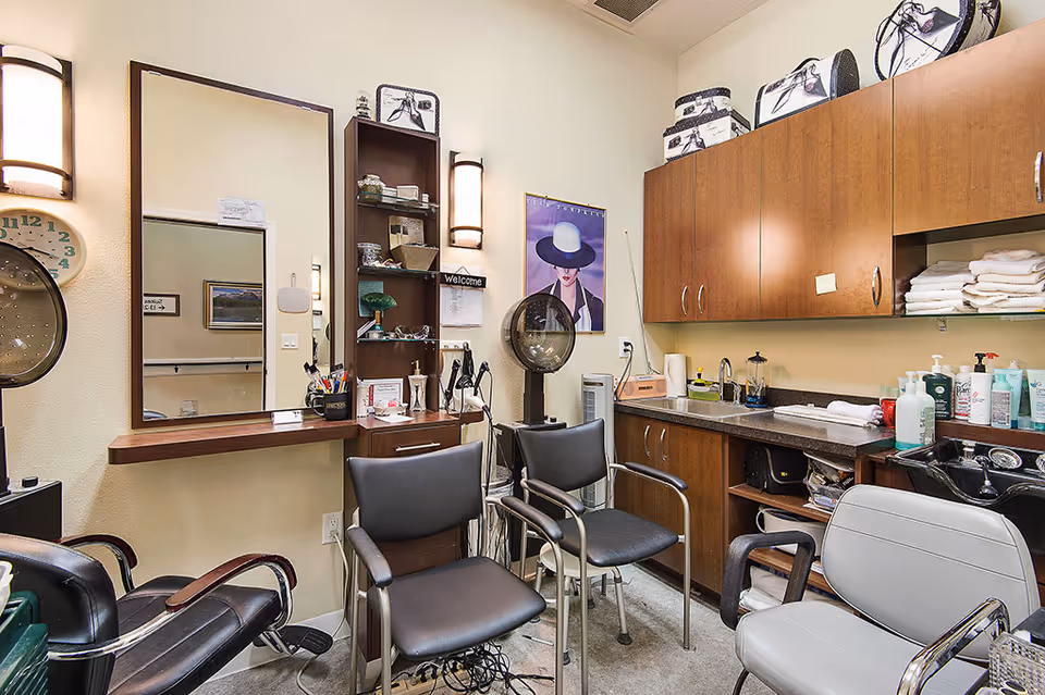 Interior view of a small salon or beauty room with multiple chairs, a large mirror, shelves with hair care products, a hair dryer, and wooden cabinets stocked with towels and various bottles.