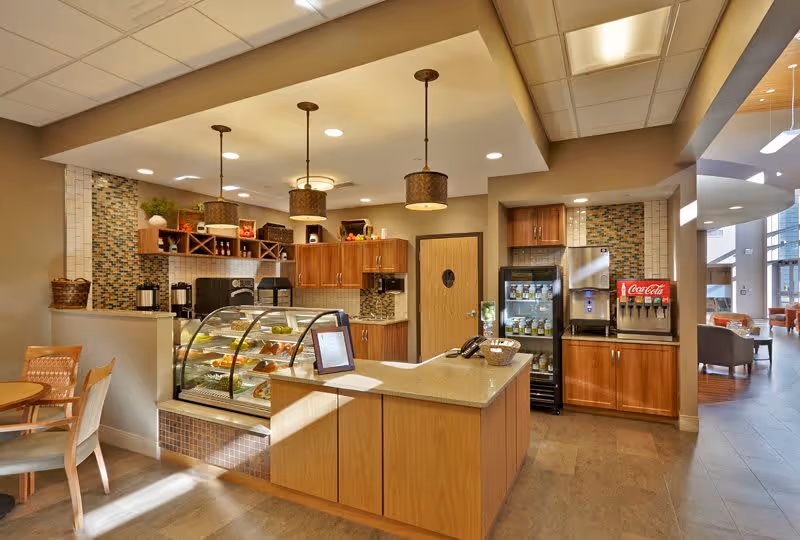 Interior view of a cafe or snack bar area within a senior living facility. The space features a counter with a glass display case containing pastries and snacks. Behind the counter are wooden cabinets, a refrigerator stocked with drinks, a Coca-Cola soda fountain machine, and coffee dispensers. Pendant lights hang from the ceiling, and there are tables and chairs for seating. The area is well-lit with natural and artificial light, and a lounge area is visible in the background.