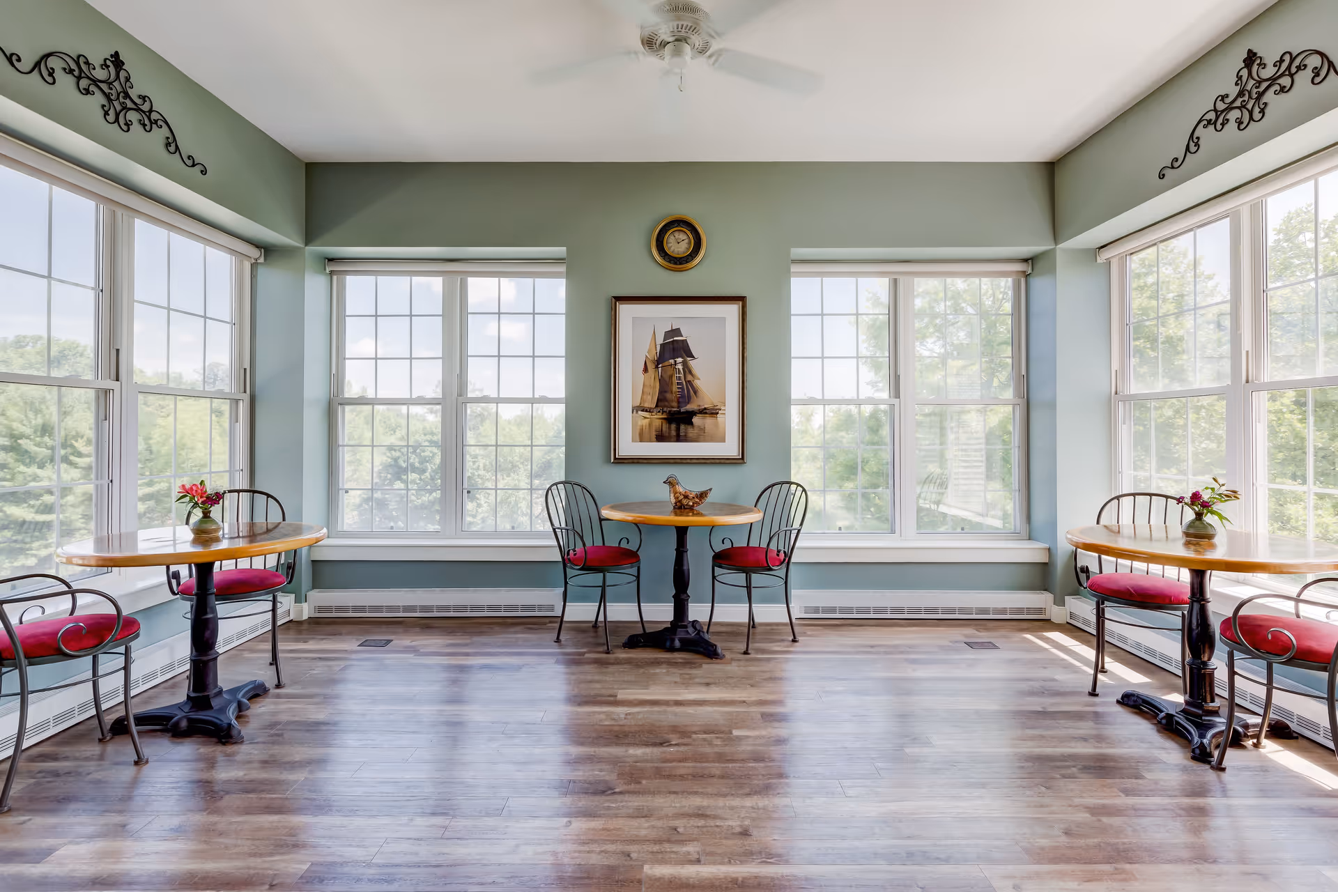 A bright room with large windows on three sides, featuring three small round wooden tables each with two metal chairs with red cushions. The walls are painted light green, and there is a framed picture of a sailing ship and a clock on the central wall. The floor is wooden, and there are small flower vases on two of the tables.