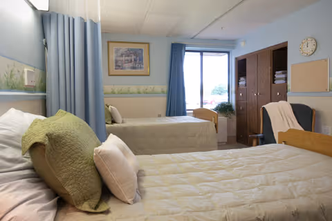 A tidy senior living bedroom with two single beds, a privacy curtain, window with blue curtains, and wooden storage cabinets.