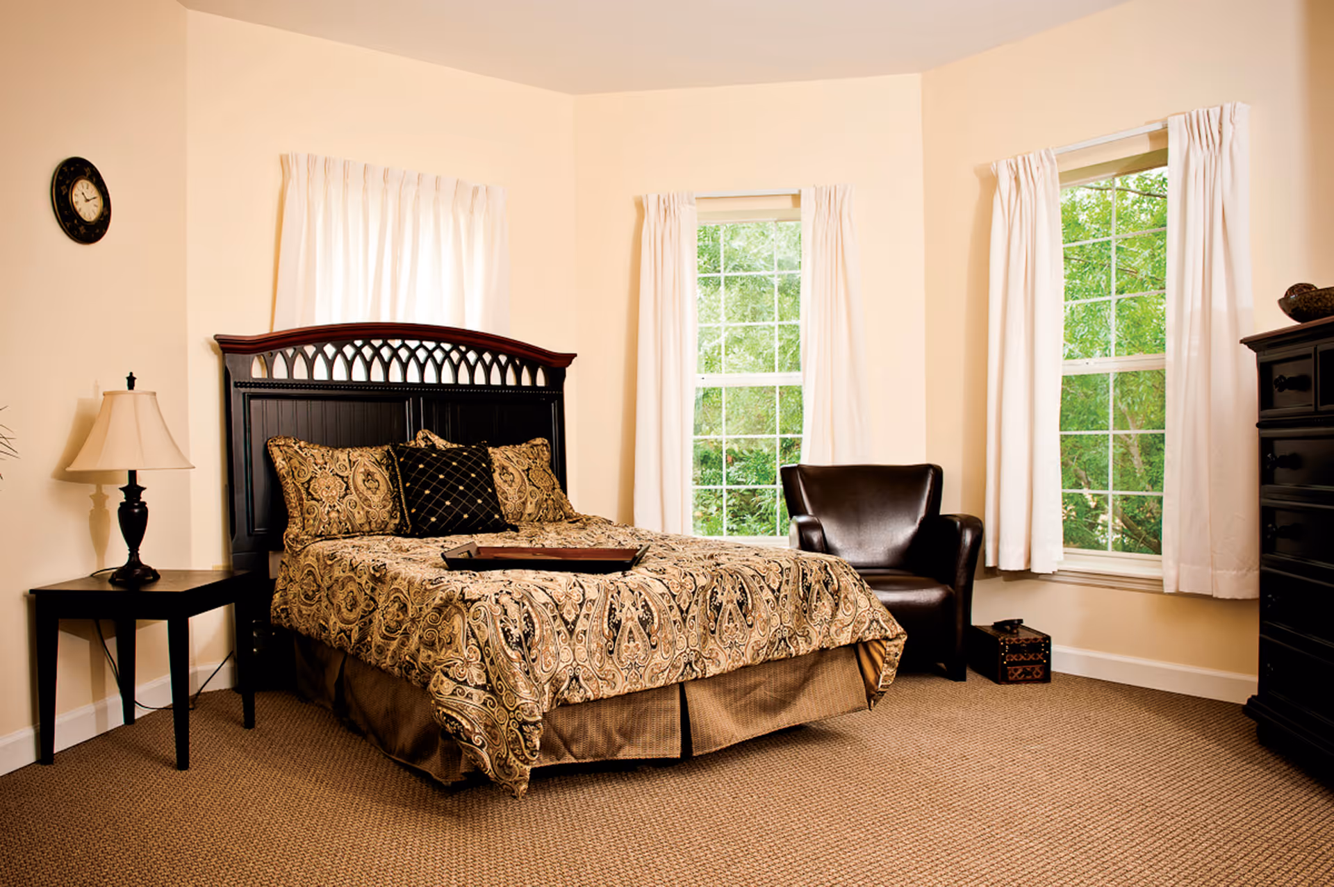 Well-lit bedroom with a patterned bed and dark headboard, a leather armchair, nightstand with lamp, and three windows with white curtains.