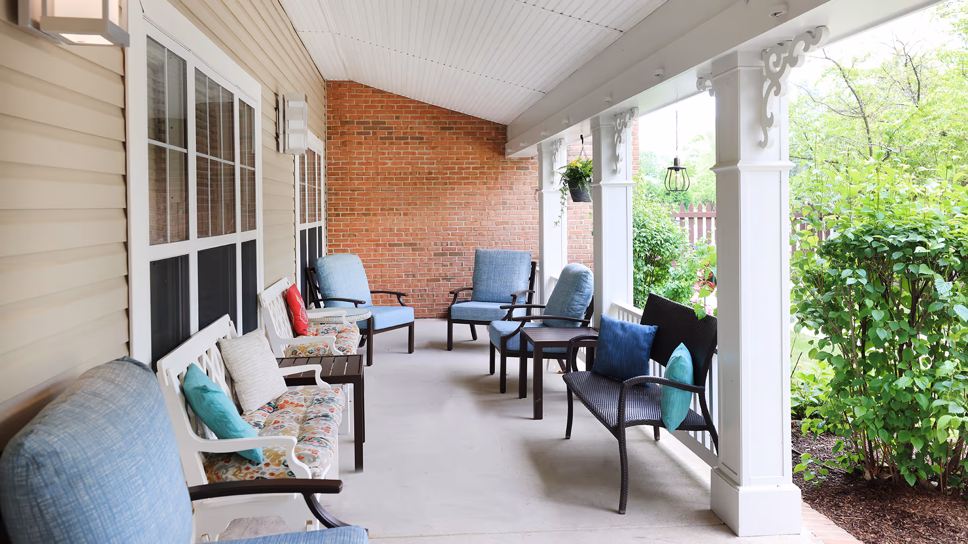 Covered outdoor porch area with multiple cushioned chairs and benches arranged along the wall and open side. The porch has white pillars with decorative brackets, a brick wall at the far end, and greenery visible outside the porch area.