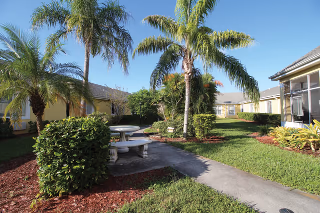 Courtyard with palm trees, grass, a concrete walkway and a round stone picnic table between single-story yellow buildings under a clear blue sky.