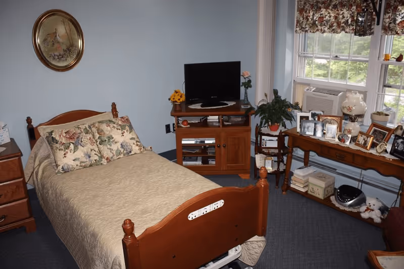 A cozy bedroom with a single wooden bed covered with a beige quilt and floral pillows. There is a wooden nightstand with a tissue box on the left, a wooden TV stand with a flat-screen TV and decorative items in the center, and a wooden table under a window on the right displaying numerous framed photos, a lamp, plants, and other small decorations. The room has light blue walls and a carpeted floor.