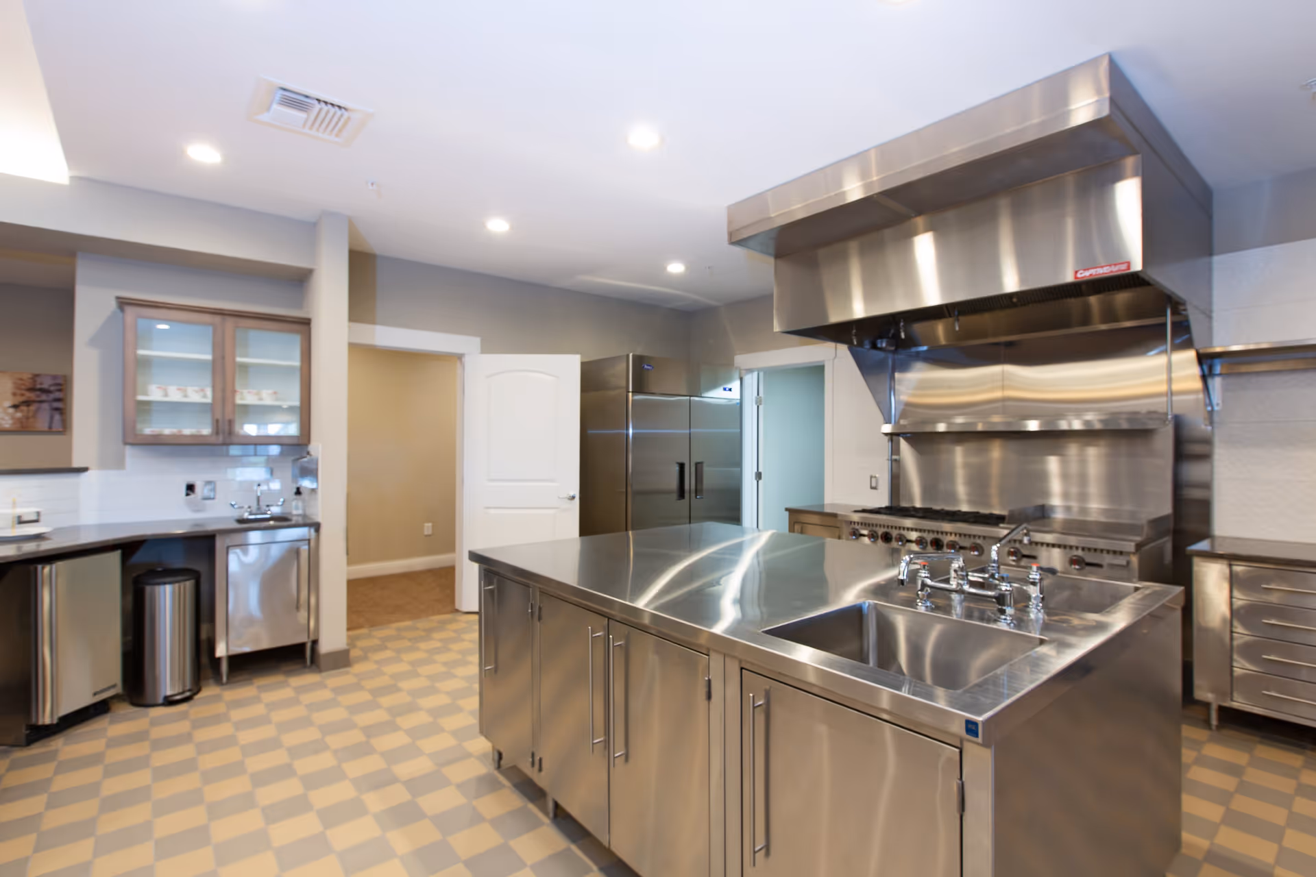 A clean, modern commercial kitchen with stainless steel appliances and fixtures. The kitchen features a large central island with a sink, a professional-grade stove with an overhead vent hood, a double-door refrigerator, and cabinets with glass doors. The floor has a checkered tile pattern, and the walls are painted in neutral tones.