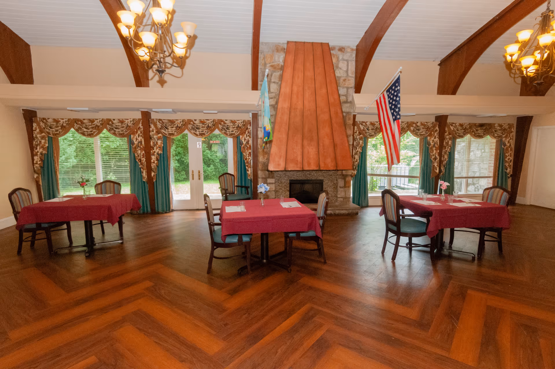 Spacious dining room with several tables covered in red tablecloths, a central stone fireplace, chandeliers, and large windows with draped curtains.