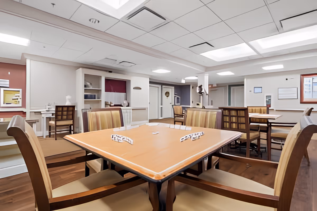 Communal dining and activity room with wooden tables and upholstered chairs arranged under recessed ceiling lights.
