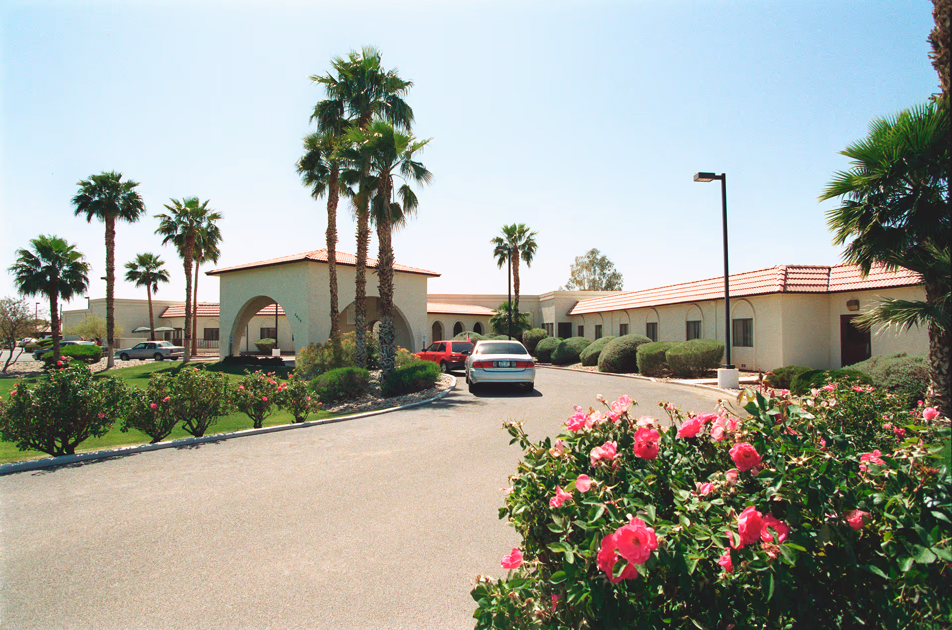 Exterior view of Life Care Center of Yuma showing a single-story building with a tiled roof, surrounded by palm trees and flowering bushes. A driveway leads to the entrance where several cars are parked under a covered archway.