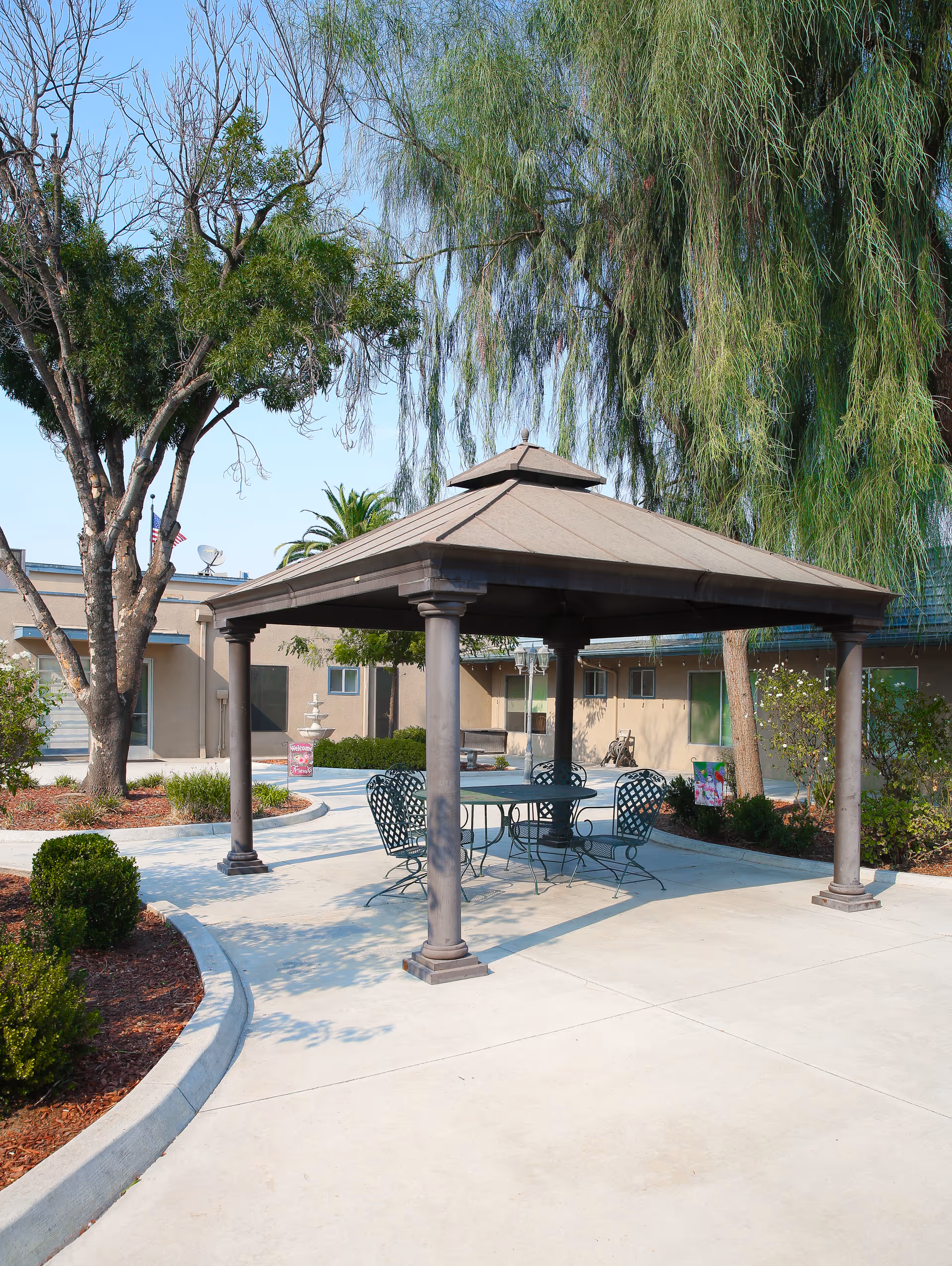 A gazebo with a metal table and chairs in a paved, landscaped courtyard surrounded by trees and low buildings.