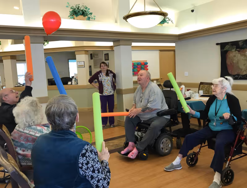Elderly residents and a staff member in a common room playfully hit a red balloon with colorful foam pool noodles.