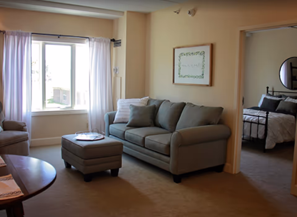 A cozy living room with a gray sofa, matching ottoman, and an armchair near a large window with white curtains. A framed picture hangs on the wall above the sofa. Through an open doorway, a bedroom with a bed, pillows, and a round mirror on the wall is visible.