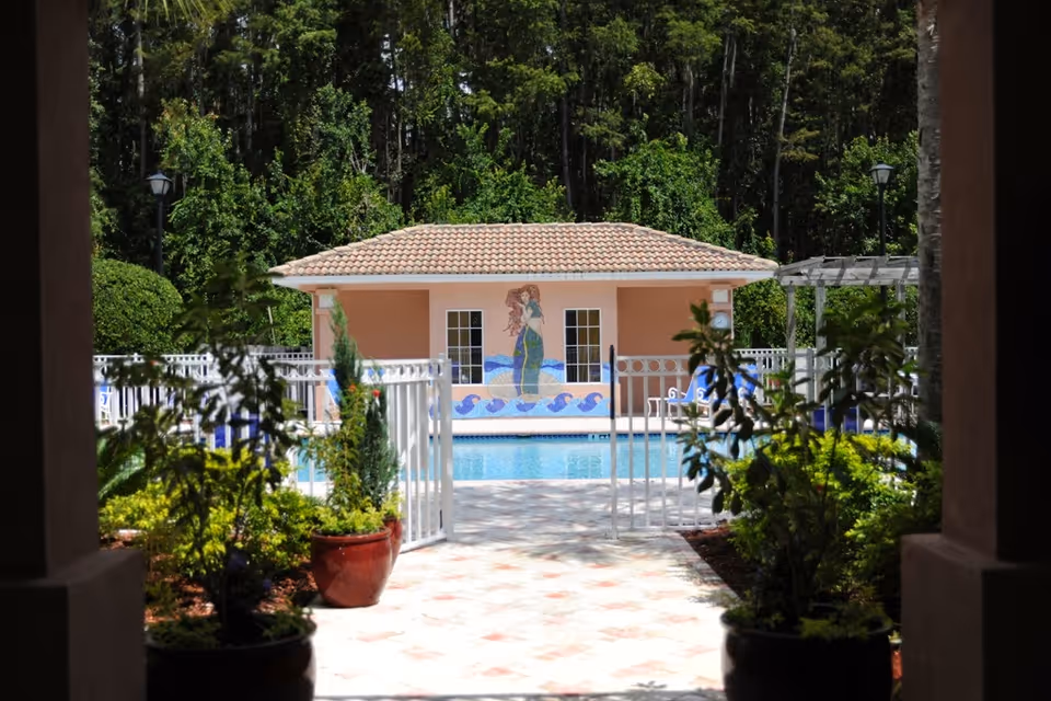 View through a shaded archway of a gated outdoor swimming pool area with a small building in the background featuring a mermaid mural. The pool area is surrounded by greenery and trees, with potted plants and lounge chairs visible.