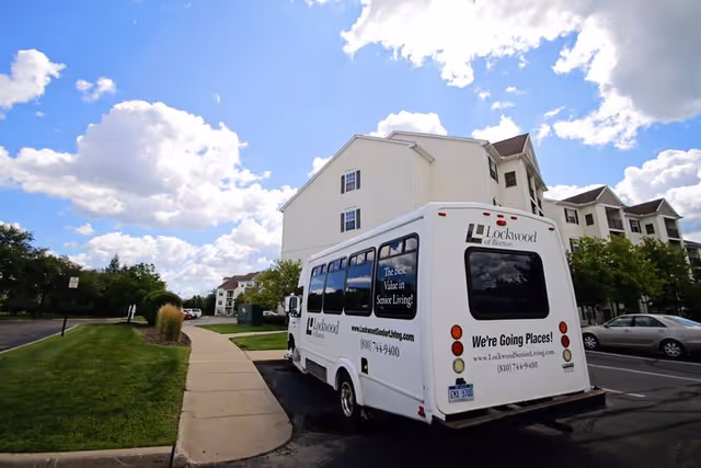 A white shuttle bus parked in a parking lot next to a sidewalk and green lawn, with a multi-story residential building in the background under a partly cloudy blue sky. The bus has signage for Lockwood of Burton senior living facility, including the phrases 'The Best Value in Senior Living!' and 'We're Going Places!' along with a phone number and website.