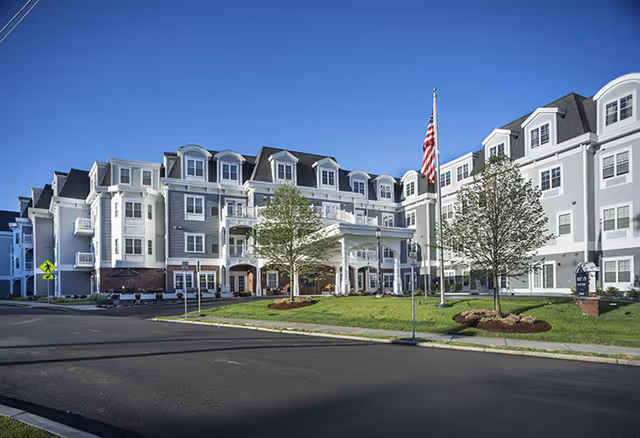 Exterior view of a large, multi-story senior living facility building with white and gray siding, multiple windows, and a covered entrance. There are two trees and a flagpole with an American flag in front of the building, along with a well-maintained lawn and clear blue sky.