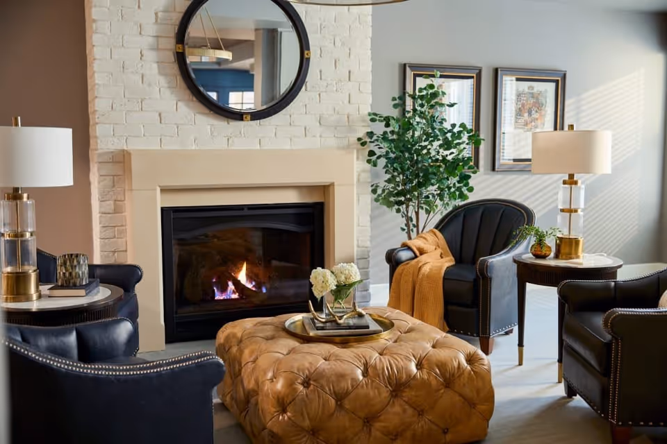 A cozy living room with a lit fireplace set in a white brick wall. Above the fireplace is a round mirror. The room features four black leather armchairs with nailhead trim arranged around a large tufted brown leather ottoman. Two side tables with modern lamps flank the chairs. A green potted plant and framed artwork decorate the wall, and sunlight streams through a window with blinds.