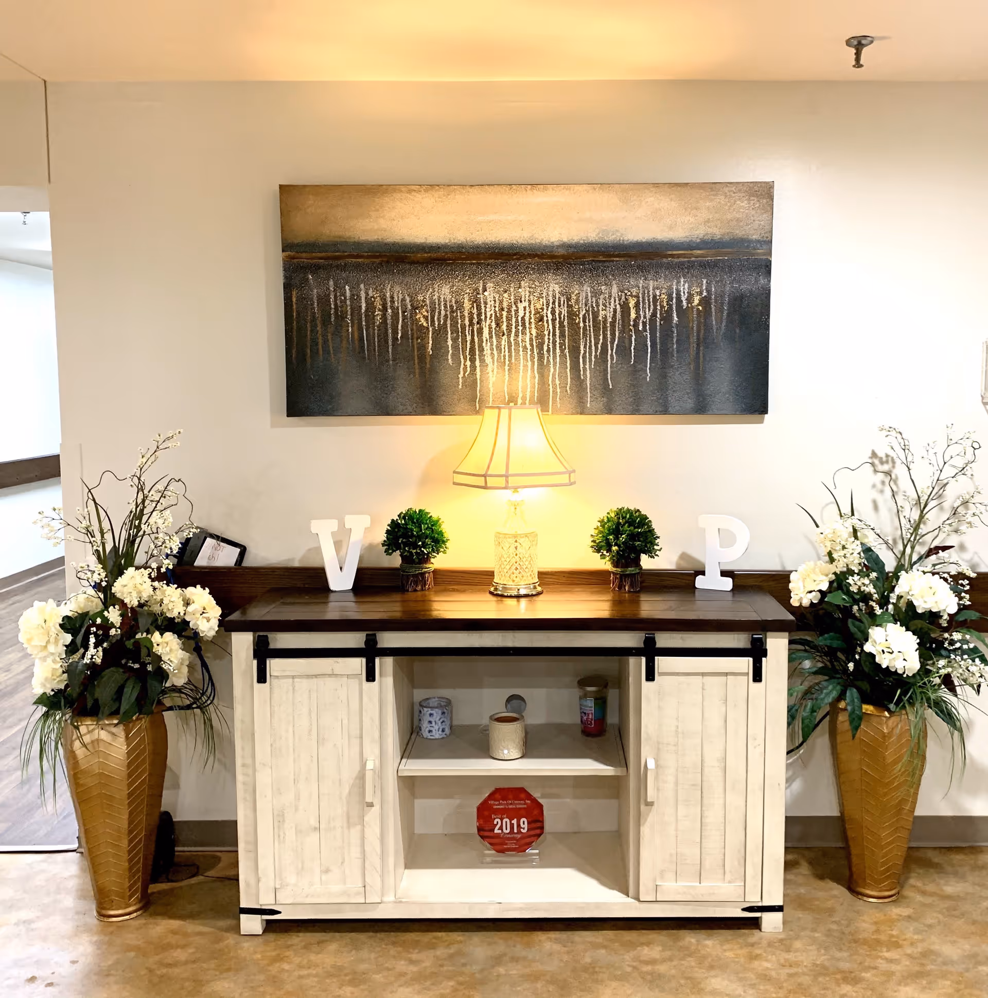 Console table with a lit lamp, artwork above, and floral arrangements on either side in a hallway.