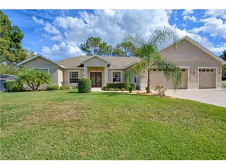 Single-story residential building with a beige exterior, a three-car garage, a front door with sidelights, and a well-maintained lawn with palm trees and shrubs under a partly cloudy sky.