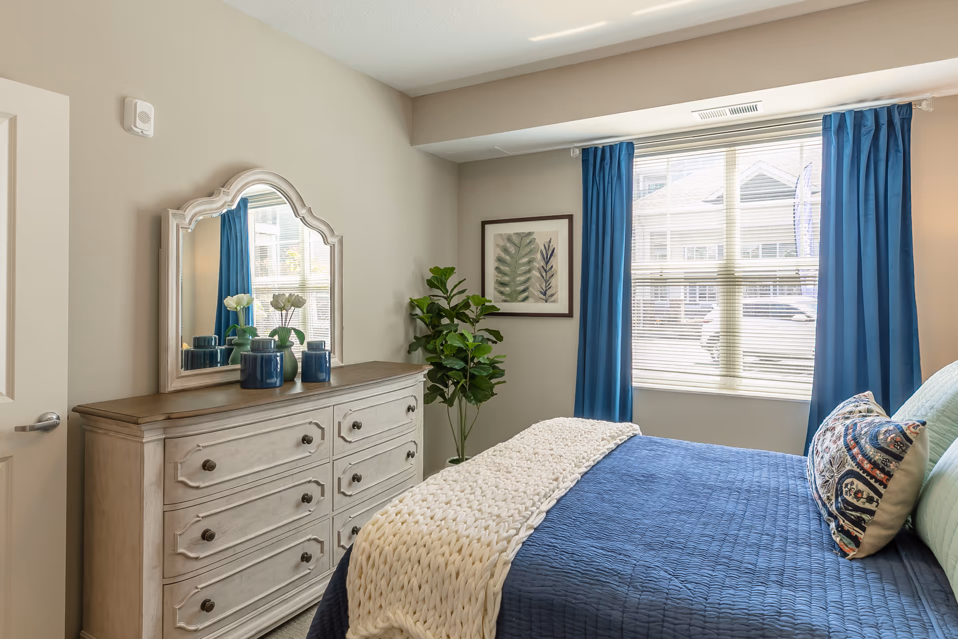 A cozy bedroom with a bed covered in a blue quilt and a white knitted throw blanket. There are decorative pillows on the bed. A wooden dresser with a large mirror on top is against one wall, adorned with blue vases and a small flower arrangement. A green potted plant is in the corner near a window with blue curtains, letting in natural light.