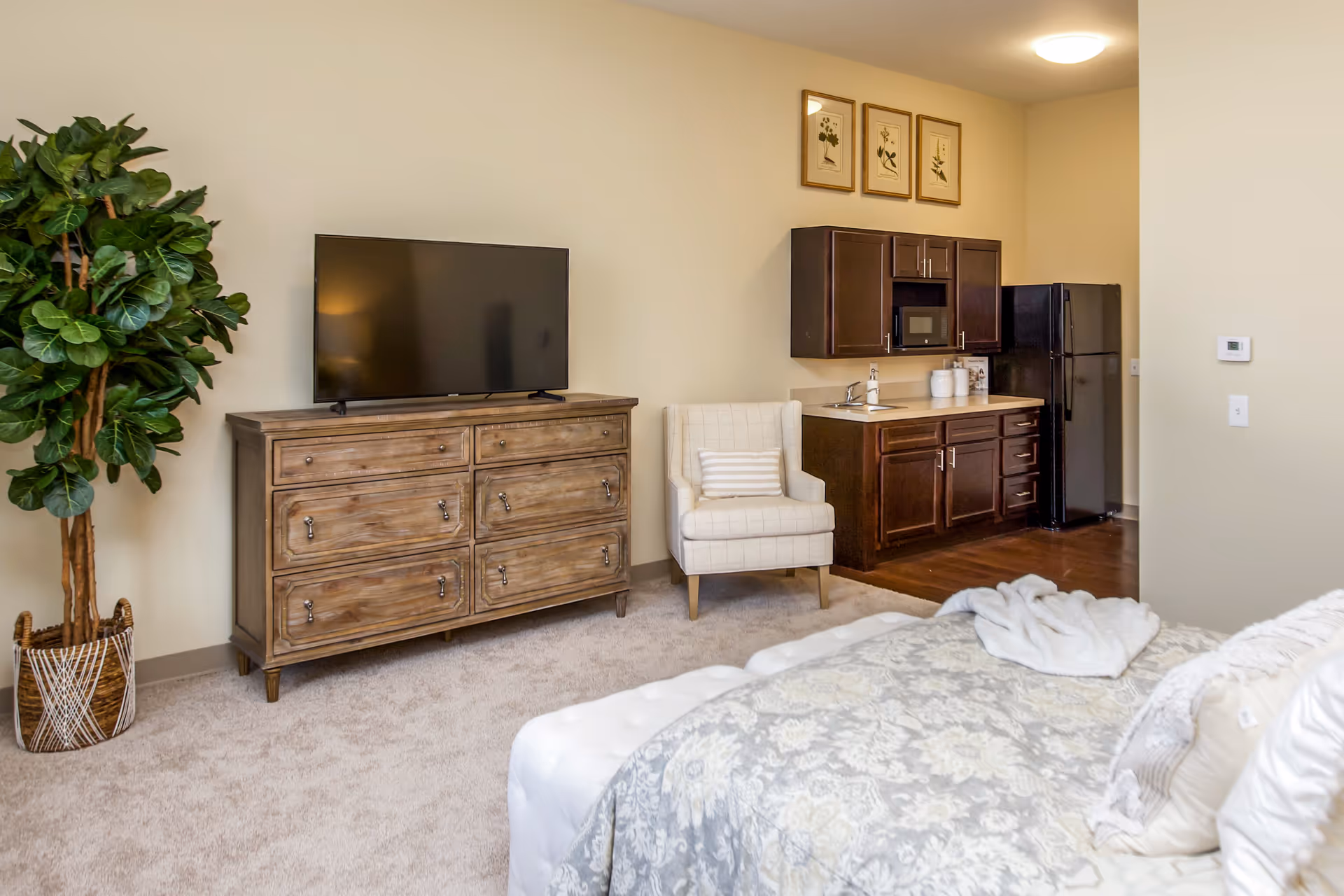 Studio apartment interior showing a bed in the foreground, a dresser with TV, an armchair, and a small kitchenette with fridge.