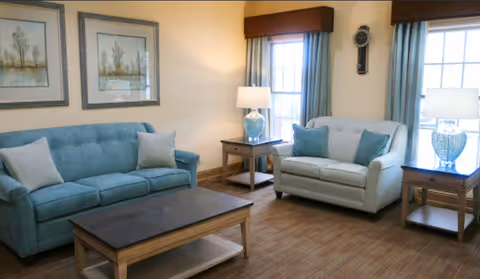 A cozy living room with two sofas, one blue and one light gray, each with matching throw pillows. There are two wooden side tables with blue and white table lamps on either side of the sofas. The room has two windows with blue curtains and brown valances, and two framed landscape paintings on the wall above the blue sofa. The floor is carpeted in a neutral tone.