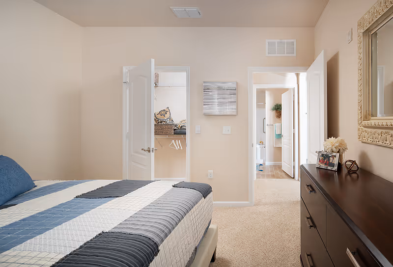 A bedroom with a bed covered in a blue, gray, and white striped quilt. The room has beige walls and carpet. There is an open door leading to a walk-in closet with shelves and hangers, and another open door leading to a bathroom. A dark wooden dresser with a framed photo, decorative items, and a large ornate mirror is on the right side of the room.