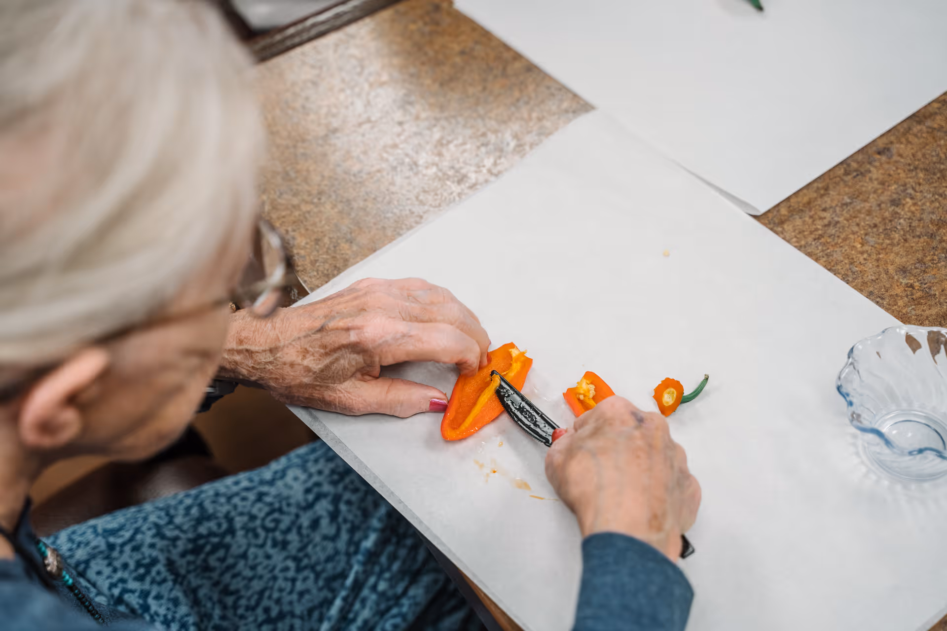 An elderly person with gray hair and glasses is slicing an orange bell pepper on a white surface using a small knife. A clear glass bowl is placed nearby on the table.