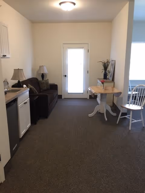 Interior view of a small living area in a retirement facility featuring a dark brown couch with two pillows, two table lamps on either side, a small kitchenette with a sink and dishwasher on the left, a white door with a window at the center back wall, and a small wooden table with a white chair on the right side near a window.