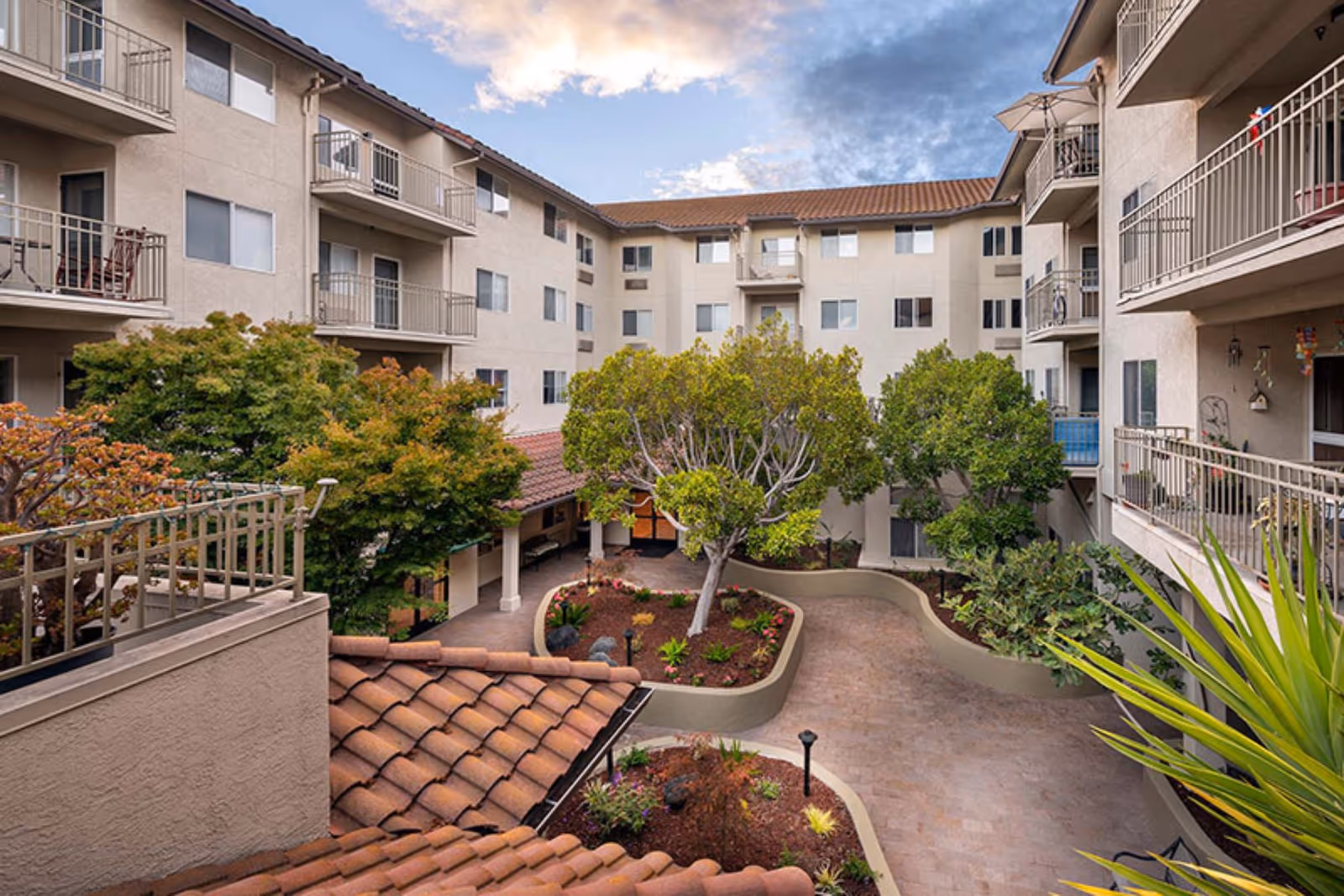 View of a multi-story senior living facility courtyard with landscaped garden beds, trees, and paved walkways surrounded by beige buildings with balconies under a partly cloudy sky.