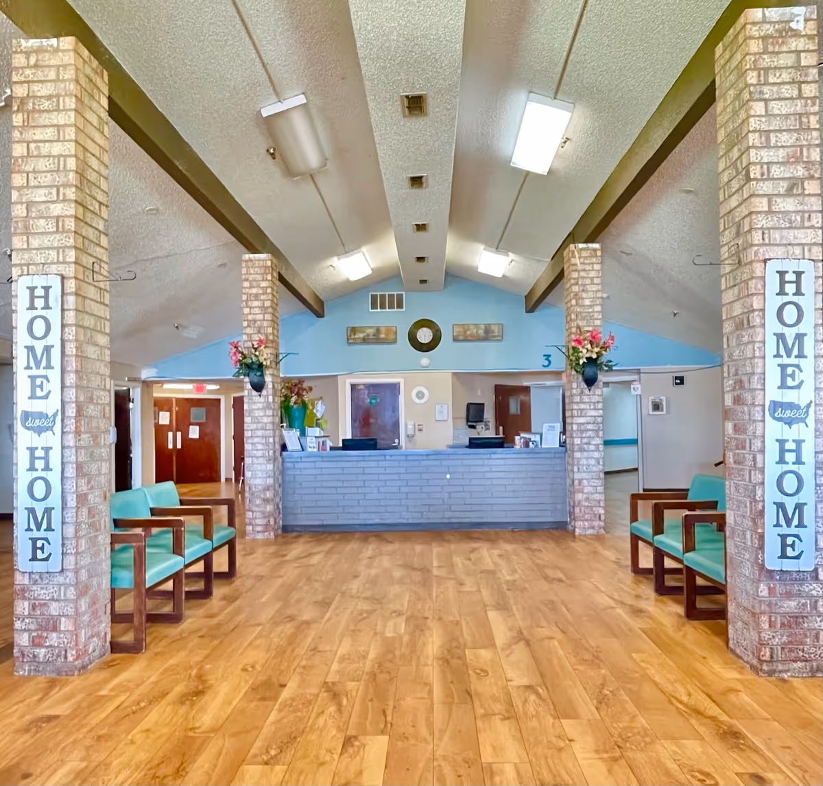 Interior view of a nursing and rehabilitation facility reception area with a wooden floor, brick pillars, and a front desk. There are green cushioned chairs along the walls and decorative signs on the pillars that say 'HOME sweet HOME'. The ceiling has fluorescent lights and the walls are painted light blue and beige.