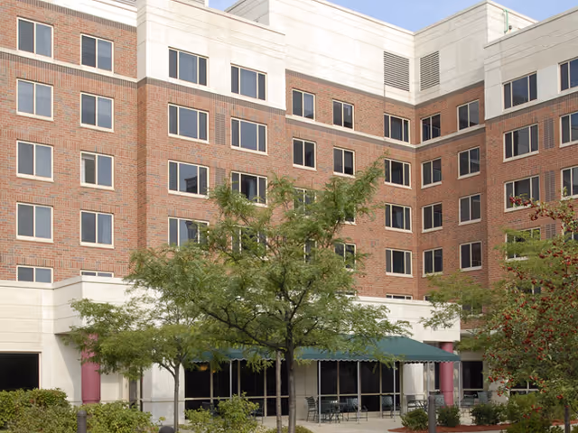 Exterior view of a multi-story senior living facility building with a brick and light-colored facade. There are several windows, trees, and shrubs in front of the building, along with a covered outdoor seating area with tables and chairs.