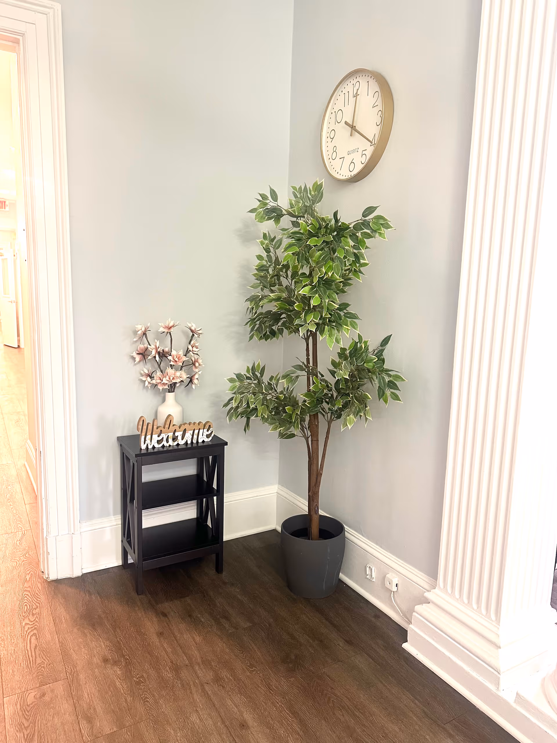 A corner of a room with light gray walls and dark wood flooring. There is a tall potted artificial plant with green leaves next to a small black shelf holding a white vase with decorative flowers and a wooden 'Welcome' sign. Above the plant, a round wall clock shows the time as 11:15. The corner is framed by white decorative molding and trim.