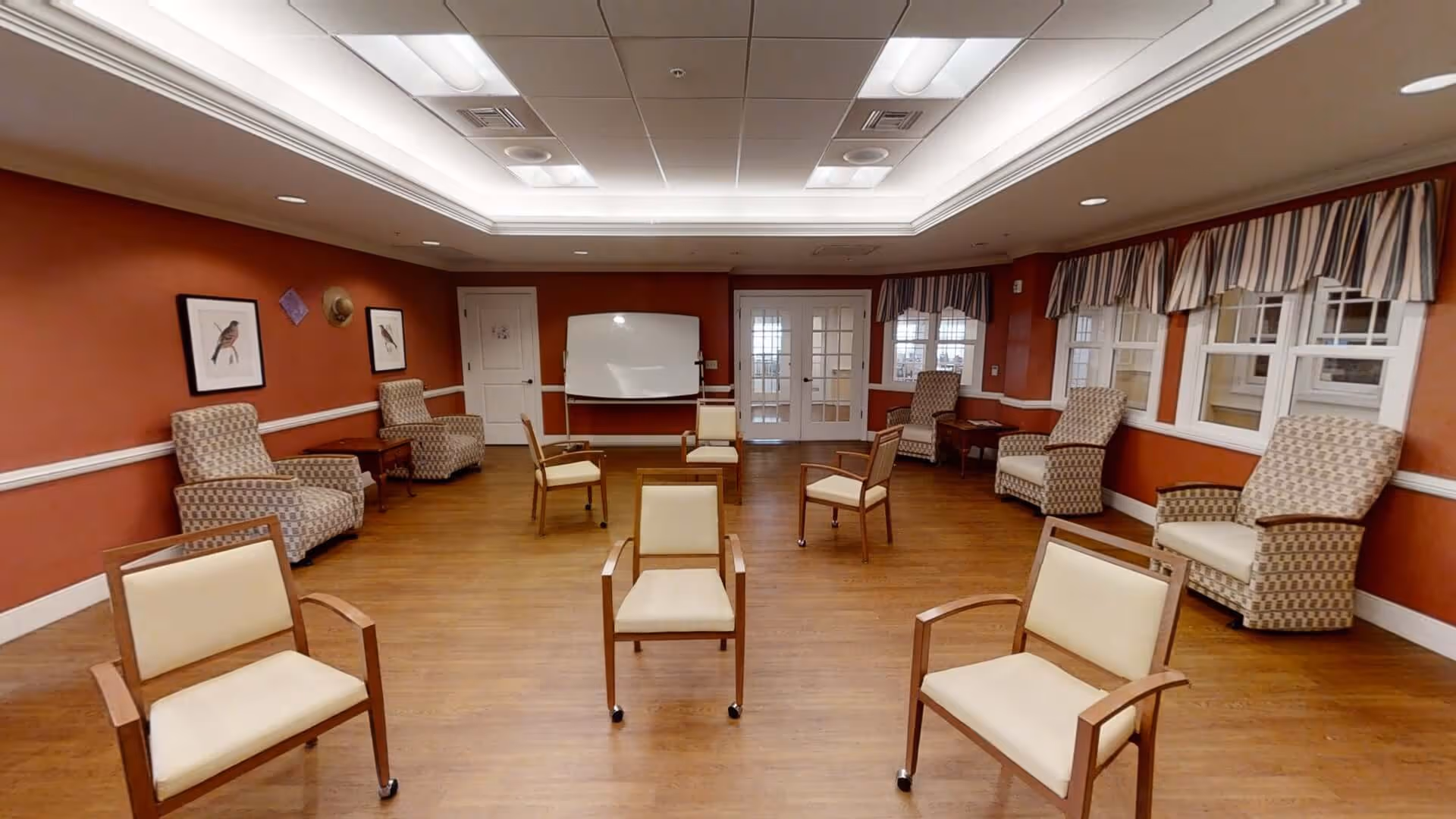 A spacious room with wooden flooring and red-orange walls, furnished with several cushioned chairs arranged in a circle and along the walls. The room features a whiteboard on a stand at the far end, framed bird artwork on the walls, and windows with striped valances. The ceiling has recessed lighting and a tray design.
