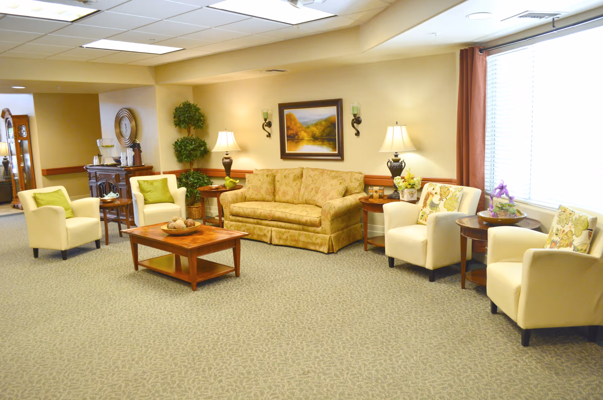 A cozy living room area in an assisted living facility featuring a patterned beige sofa, four cream-colored armchairs with decorative pillows, two wooden side tables with lamps, a wooden coffee table with a decorative bowl, a large window with blinds and curtains, a framed landscape painting on the wall, and some green plants in the corner.