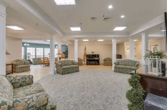 A spacious living room area in a senior living facility with floral patterned sofas arranged around a large carpeted floor. The room features white columns, a piano against the far wall with an American flag beside it, and large windows letting in natural light. There are also lamps and a small table with decorative plants.