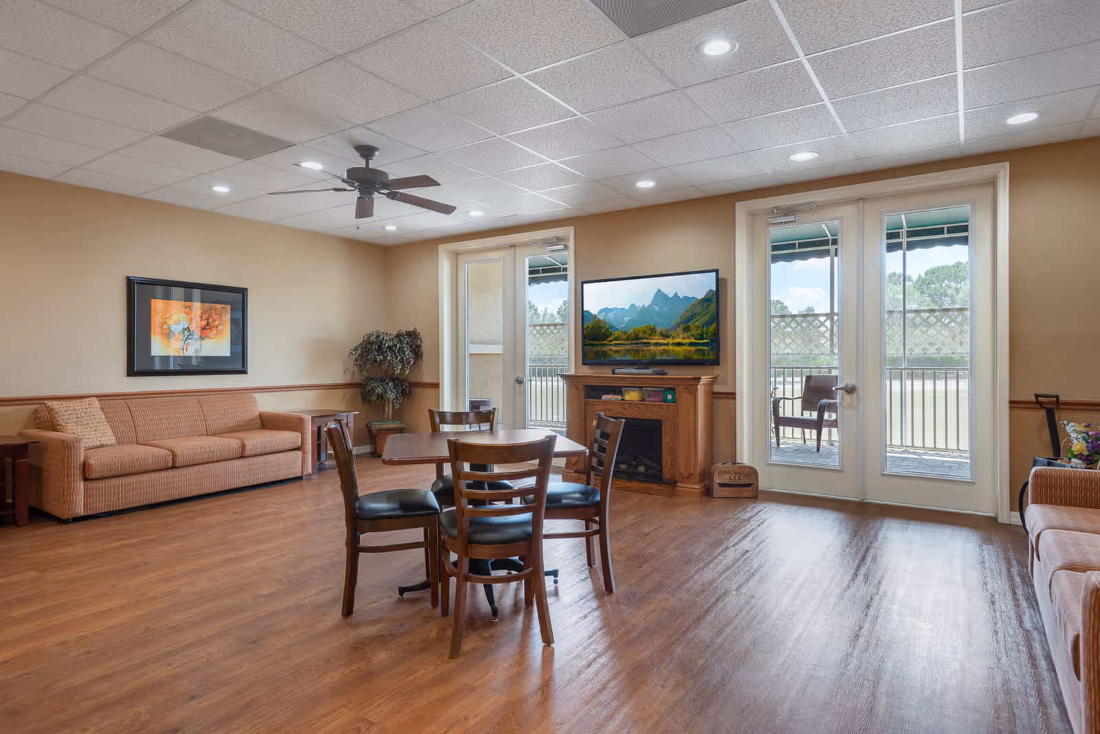 A spacious living room with wooden flooring, a ceiling fan, and recessed lighting. The room features a brown sofa with a patterned cushion, a wooden table with four chairs, a flat-screen TV mounted above a wooden fireplace, and double glass doors leading to an outdoor patio with chairs. There is a framed artwork on the wall and a potted plant in the corner.
