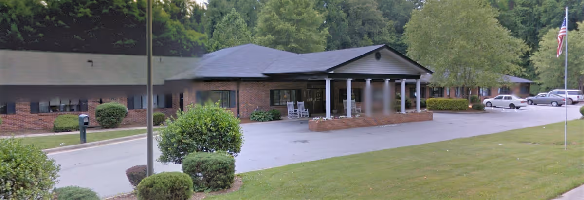 Exterior view of a single-story brick building with a covered entrance supported by white columns. There are several bushes and trees around the building, a flagpole with an American flag, and a parking area with cars. The scene is set in a green, landscaped area with a lawn in the foreground.