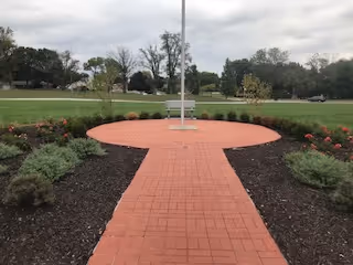 Brick walkway leading to a circular brick plaza with a flagpole and bench, surrounded by landscaped beds and lawn.