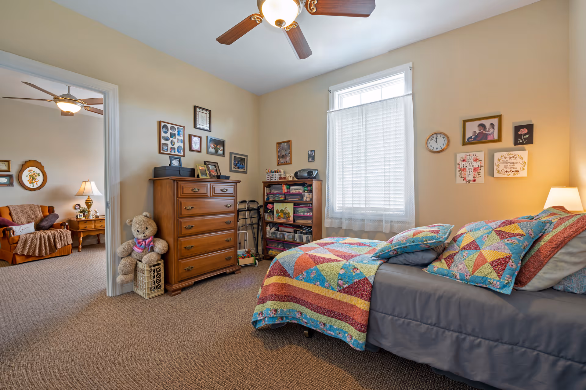 A cozy bedroom in an assisted living facility featuring a bed with a colorful patchwork quilt and matching pillows. The room has beige walls adorned with framed pictures and decorative art. A wooden dresser with photo frames and a large teddy bear sitting on a basket are visible. A window with sheer white curtains lets in natural light. An adjacent room with a sitting area and ceiling fan is partially visible through an open doorway.