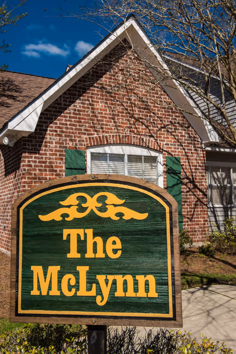 A wooden sign with green background and yellow text that reads 'The McLynn' in front of a brick building with white trim and green shutters under a clear blue sky.