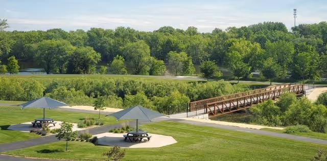 A scenic outdoor area with green grass, trees, and a wooden pedestrian bridge crossing over a small body of water. There are picnic tables with large umbrellas providing shade on concrete pads in the grassy area.