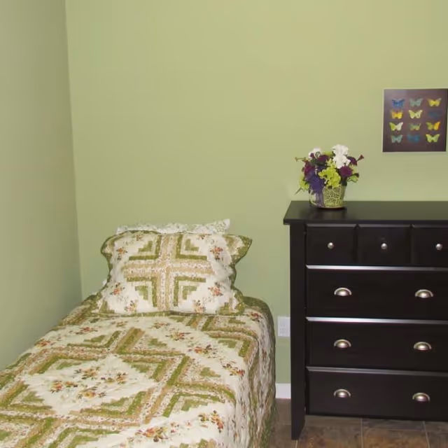 A small bedroom with a single bed covered in a green and beige patterned quilt and matching pillow. Next to the bed is a dark wooden dresser with silver handles, topped with a vase of colorful flowers. A framed picture of butterflies hangs on the light green wall above the dresser.