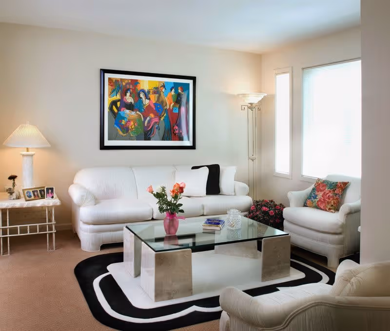 A bright and cozy living room with white upholstered furniture including a sofa, armchair, and another chair partially visible. A glass coffee table with a pink vase holding flowers and some books is centered on a black and white patterned rug. The walls are light-colored with a colorful framed painting above the sofa. A side table with a lamp and framed photos is on the left, and a tall floor lamp is in the corner near two windows with blinds.